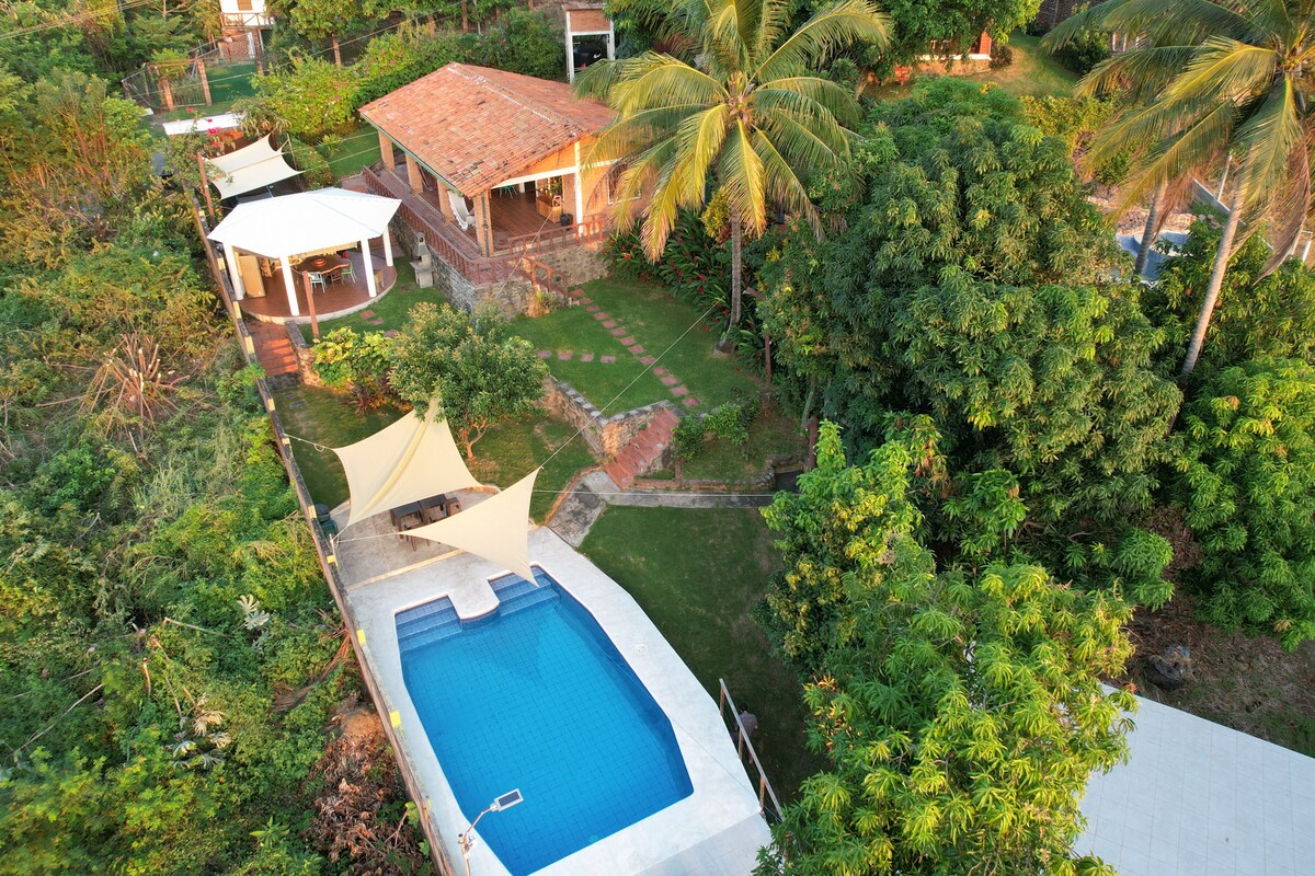 An aerial view captures the beach house surrounded by lush greenery, featuring a spacious outdoor area and private pool. The property's main structure has a terracotta roof, with shaded seating areas visible. The inviting pool is framed by landscaped grass and trees.