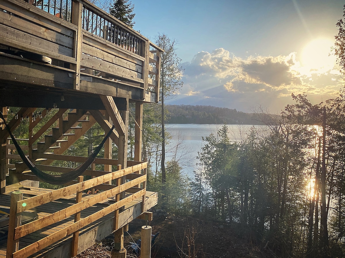 The image captures a view of the chalet’s wooden deck extending from the structure, overlooking Lake Patrick. A hammock hangs nearby, surrounded by lush trees. The setting sun casts warm light across the water, creating a serene atmosphere with soft reflections on the lake's surface.