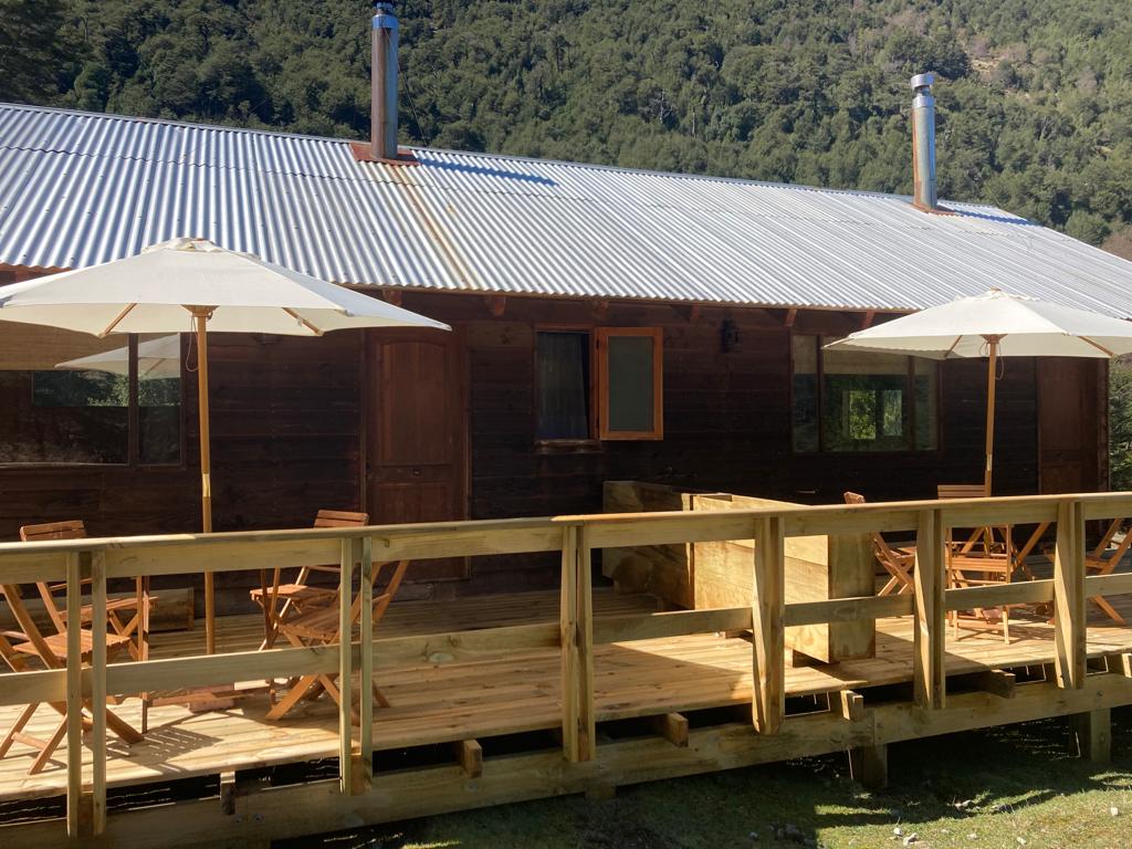 A wooden structure with a metal roof is surrounded by greenery, featuring a spacious deck. Two wooden tables with matching chairs are shaded by large umbrellas, providing an inviting outdoor seating area. Large windows allow for natural light to enter the cabin.