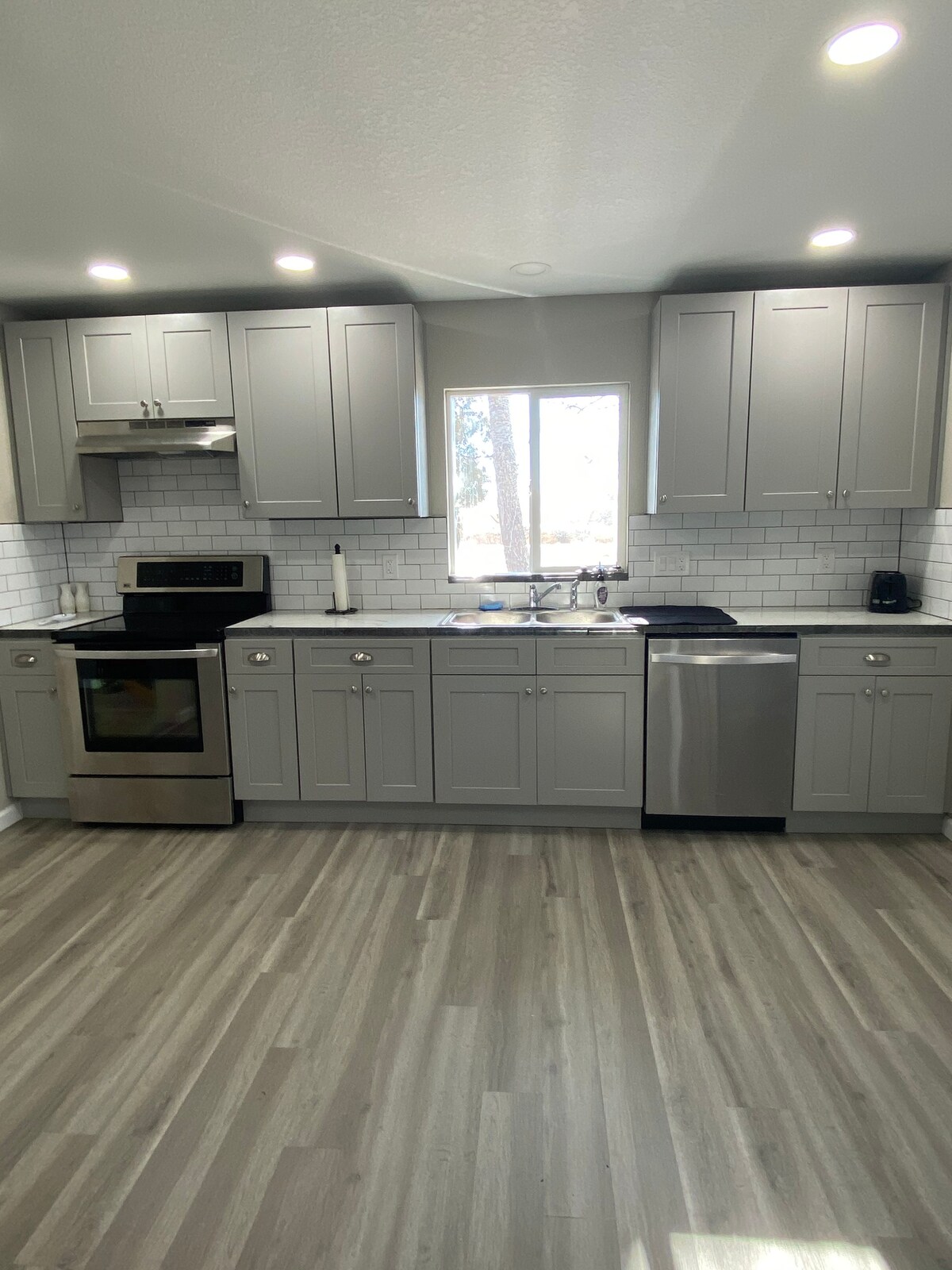 A modern kitchen features gray cabinetry with sleek hardware and stainless steel appliances. A window allows natural light to brighten the workspace, while white tile backsplash adds a clean aesthetic. Countertops provide ample space for meal preparation.