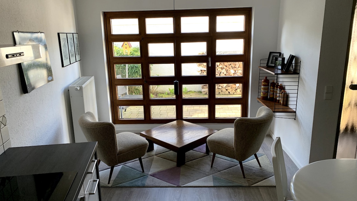 A comfortable seating area features two beige chairs positioned around a wooden coffee table. A large window with multiple panes allows natural light to illuminate the space and offers a view of the garden outside. Wall-mounted shelves display decorative items.