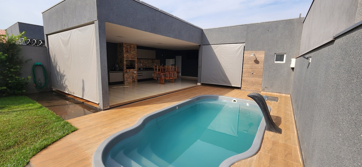 An inviting pool area is visible, featuring a sleek design complemented by a cascading waterfall feature. The surrounding space is well-maintained with green grass, and an open layout connects the pool to the leisure area, which includes a modern outdoor kitchen and dining setup.