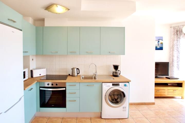 A modern kitchen space is featured, equipped with a refrigerator, microwave, and oven. Light blue cabinetry complements the wooden countertop. A washing machine is positioned beside the sink, with additional appliances including a kettle and coffee maker, providing convenience for guests.