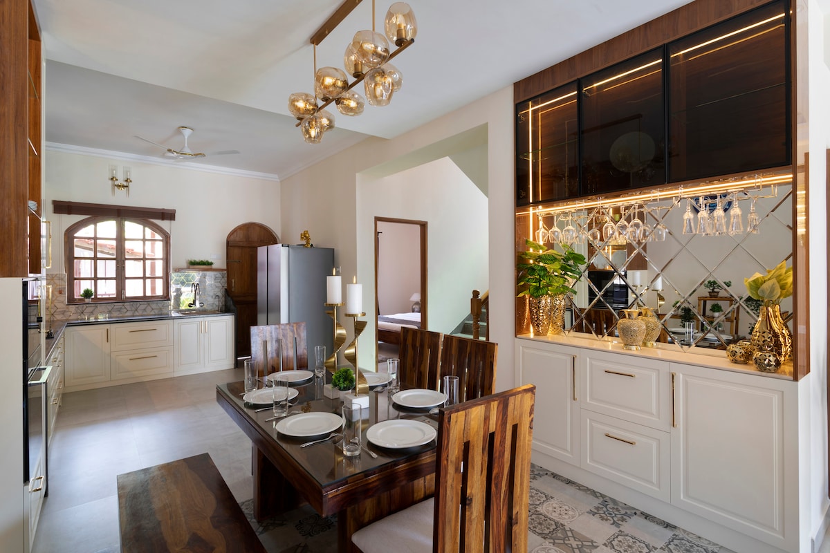 A modern kitchen and dining area features a wooden dining table set for six, complemented by stylish wooden chairs. A well-equipped kitchen showcases sleek cabinetry and a large refrigerator. Natural light enters through arched windows, creating a warm and inviting space.