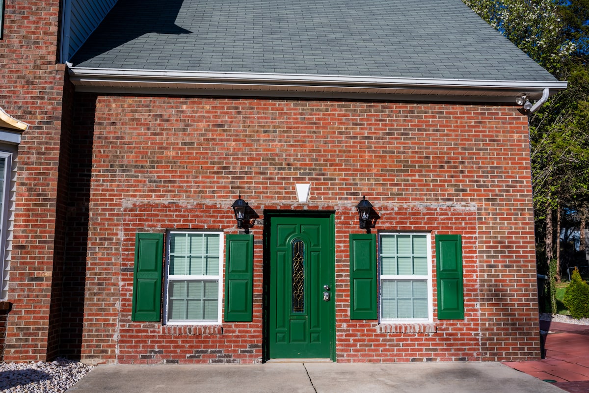 The exterior of the home features a brick façade with green shutters and a matching front door. Two windows are symmetrically placed beside the entrance, each framed by the shutters. Light fixtures are installed on either side of the door, enhancing the home's appeal.