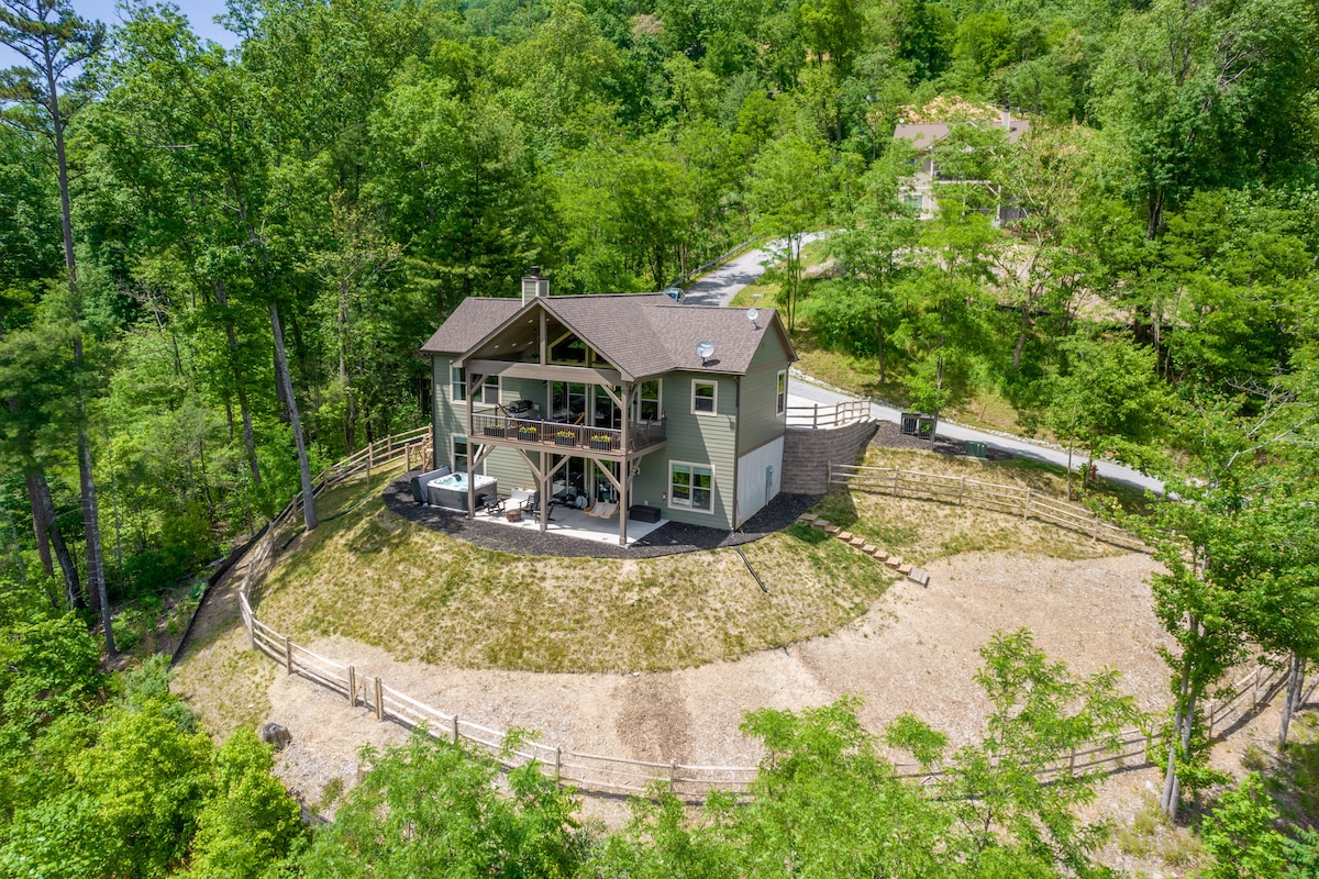 An aerial view reveals a spacious two-story cottage nestled among greenery. The circular driveway leads to the entrance, with a large deck extending from the upper level. The surrounding area features well-maintained landscaping and a natural hillside backdrop.