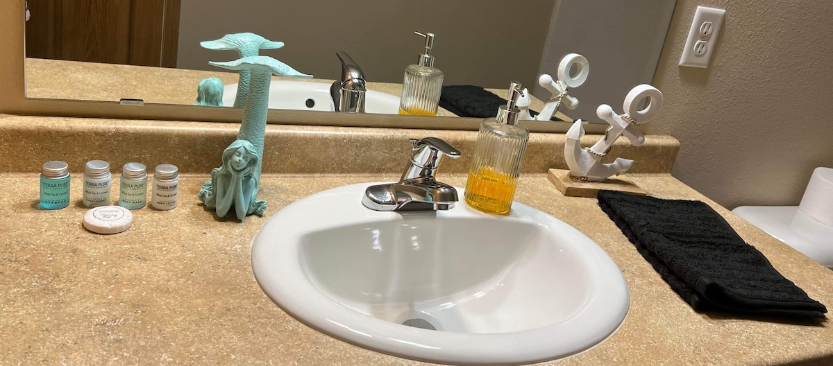 A modern bathroom sink area is presented with a round, white basin and a sleek faucet. A textured countertop displays decorative items, including a small blue sculpture, glass containers, and a dark towel. Natural light reflects off the surfaces, enhancing the clean and organized appearance.