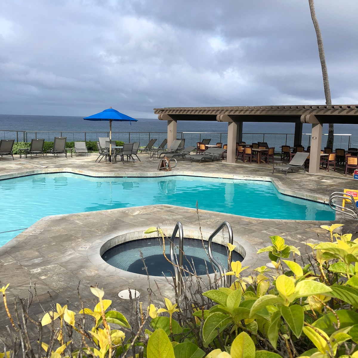A tranquil swimming pool area features a large pool with a circular hot tub nearby. Ample lounge chairs are arranged around the pool, complemented by patio umbrellas. The ocean is visible in the background under a cloudy sky, with lush greenery framing the scene.