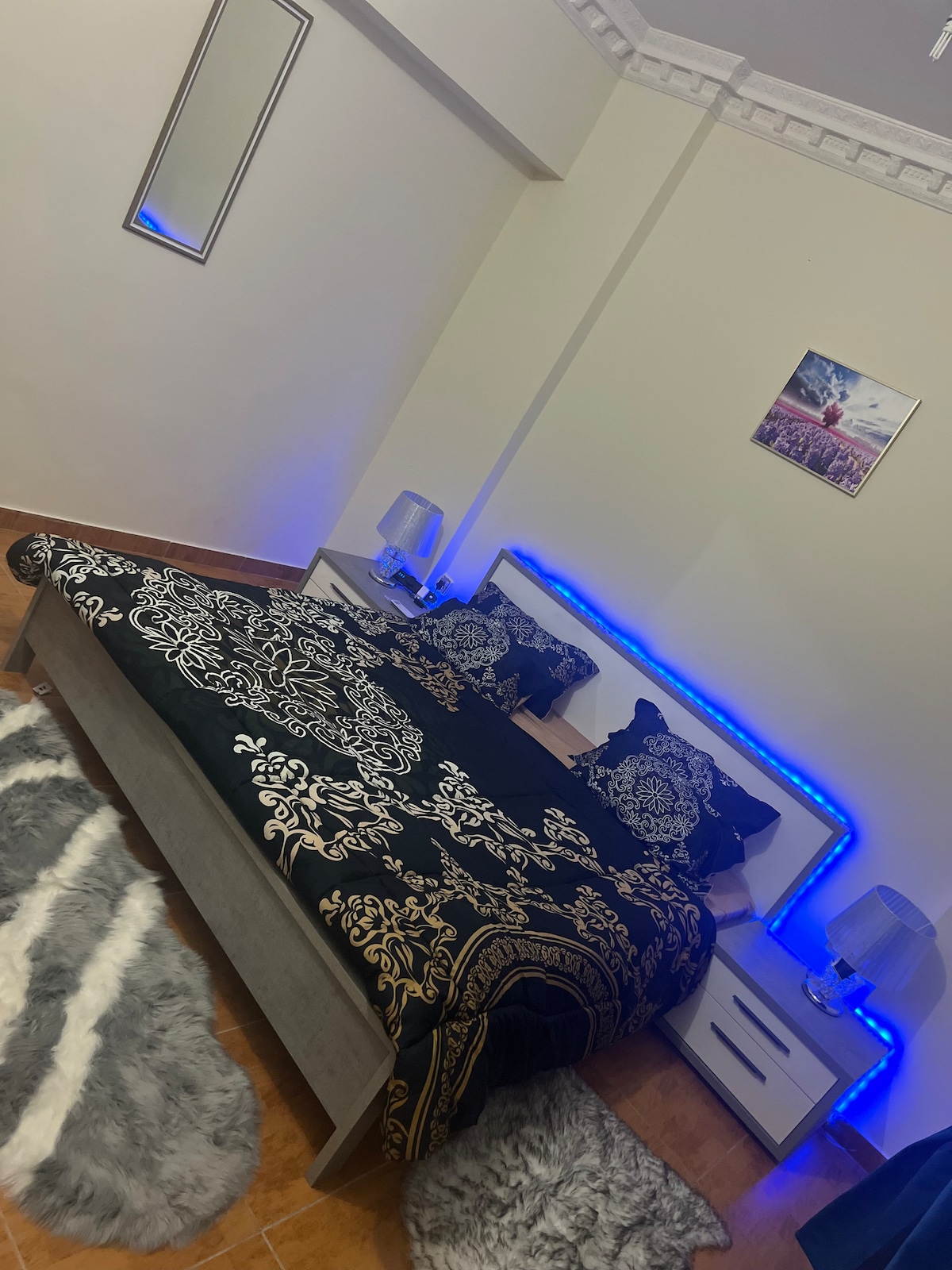 A spacious bedroom features a large bed adorned with a decorative black and gold blanket. Soft lighting around the bed enhances the ambiance. Two bedside tables with lamps flank the bed, while a framed picture adorns the wall above, and a plush rug covers part of the floor.