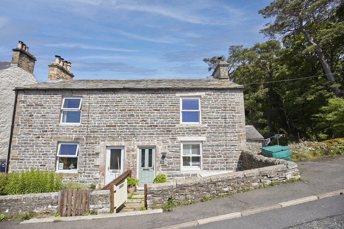 A charming stone cottage is set against a clear blue sky, showcasing two large windows that offer natural light. The entrance features a small wood railing leading to the door, while the surrounding greenery enhances the inviting atmosphere of the property.