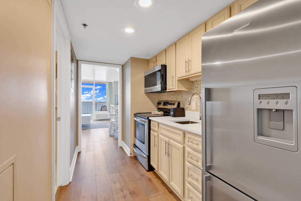 The kitchen area features light-colored cabinetry with a spacious countertop and modern appliances, including a stainless steel refrigerator. A doorway leads to a brightly lit balcony while the floor is finished with warm wood planks, providing a welcoming pathway through the space.
