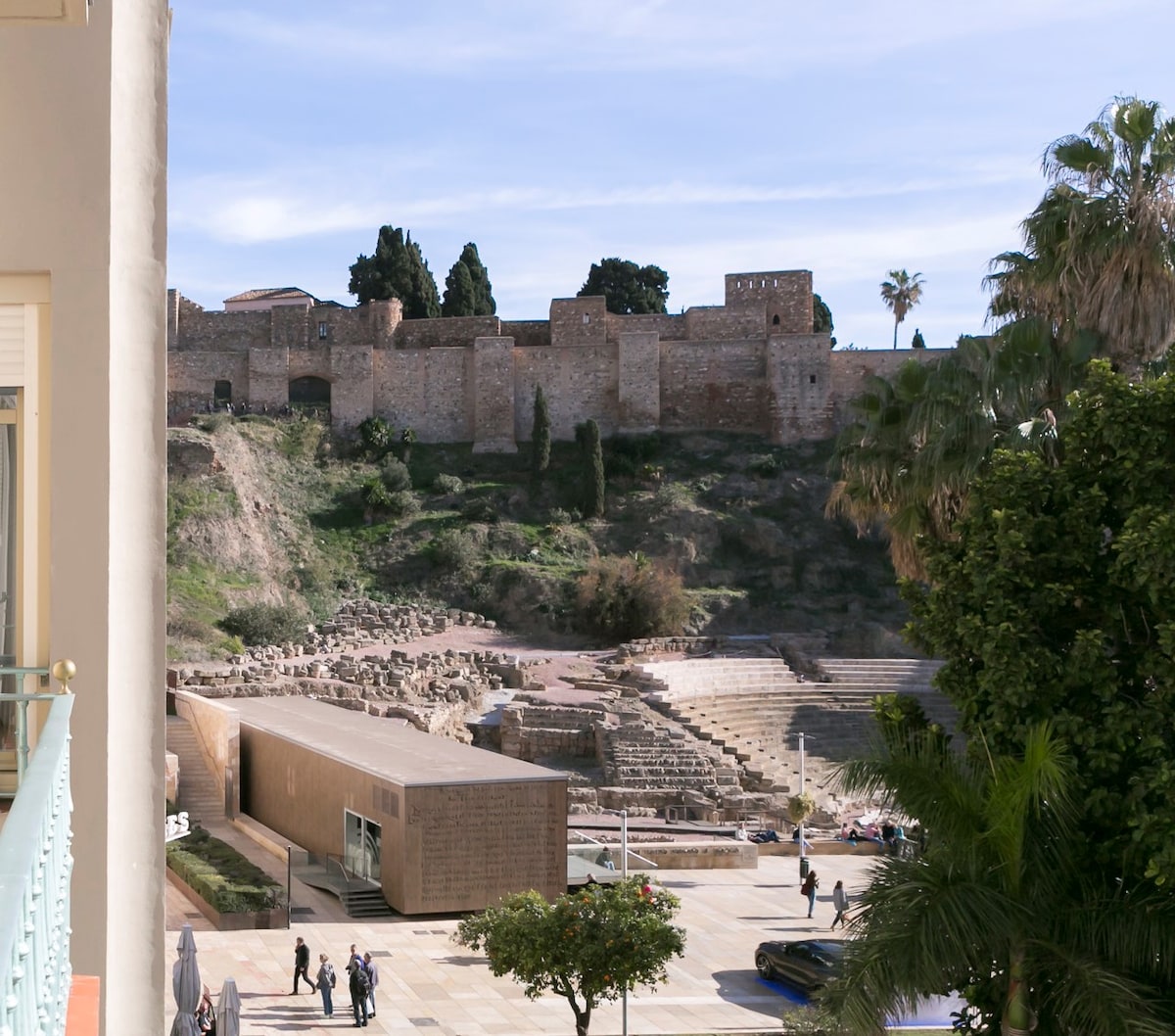 The image captures a view of the Roman Theatre of Málaga, showcasing its ancient structure alongside scenic greenery. The backdrop features the Alcazaba fortress with its stone walls. A sunlit plaza is visible in the foreground, where visitors stroll amid palms and landscaping.