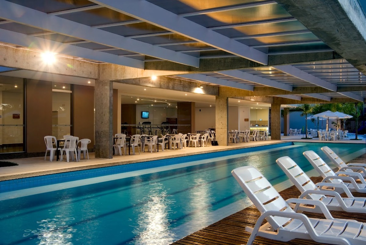 A modern pool area features a clear, inviting pool flanked by white lounge chairs. Soft lighting enhances the space, creating a relaxing atmosphere. An adjoining dining area offers tables and chairs, with indoor seating visible through expansive glass doors.
