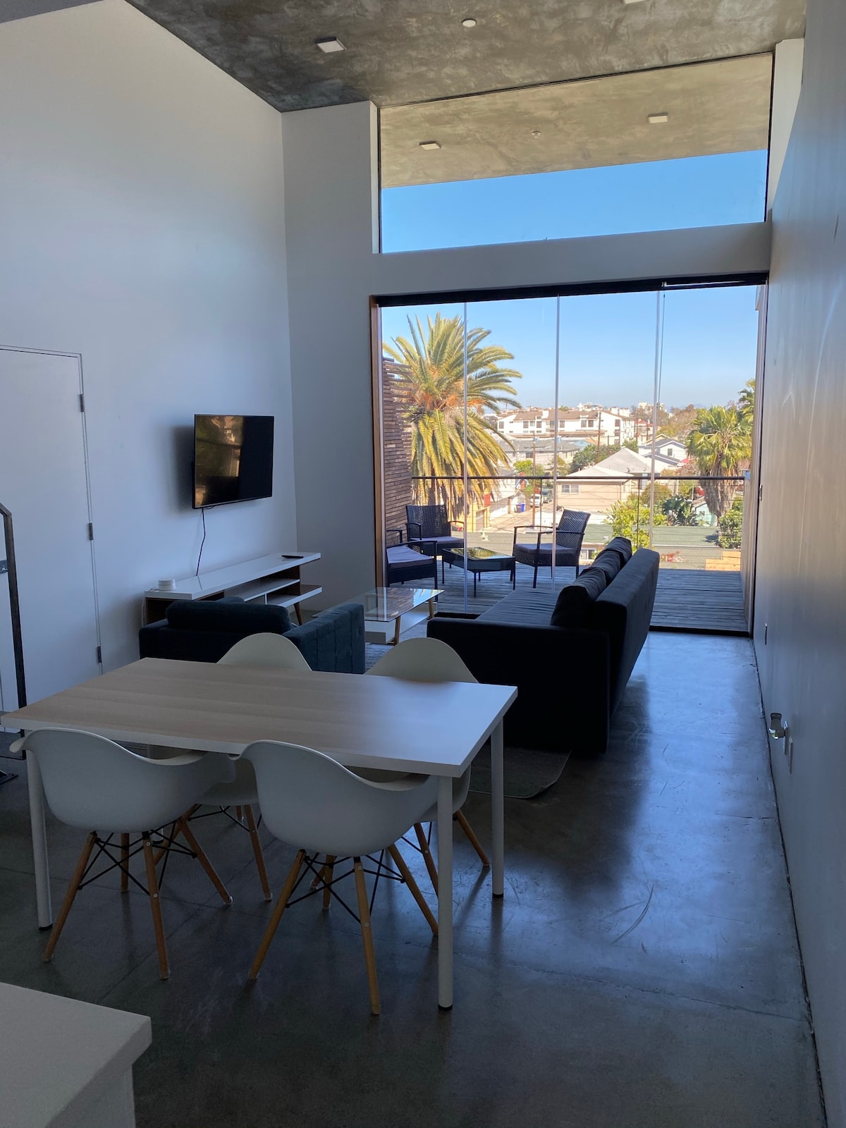 A modern living space features large glass windows providing views of palm trees and the neighborhood. A comfortable sofa and a coffee table are positioned in the seating area, while a dining table with white chairs sits nearby. A wall-mounted television is visible.