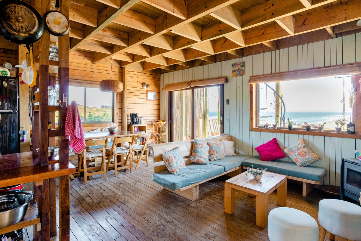 A spacious living area features wooden beams and a cozy, low seating arrangement with decorative cushions. Large windows provide natural light and offer views of the ocean. A dining table is visible in the background, accompanied by wooden chairs, creating an inviting atmosphere.