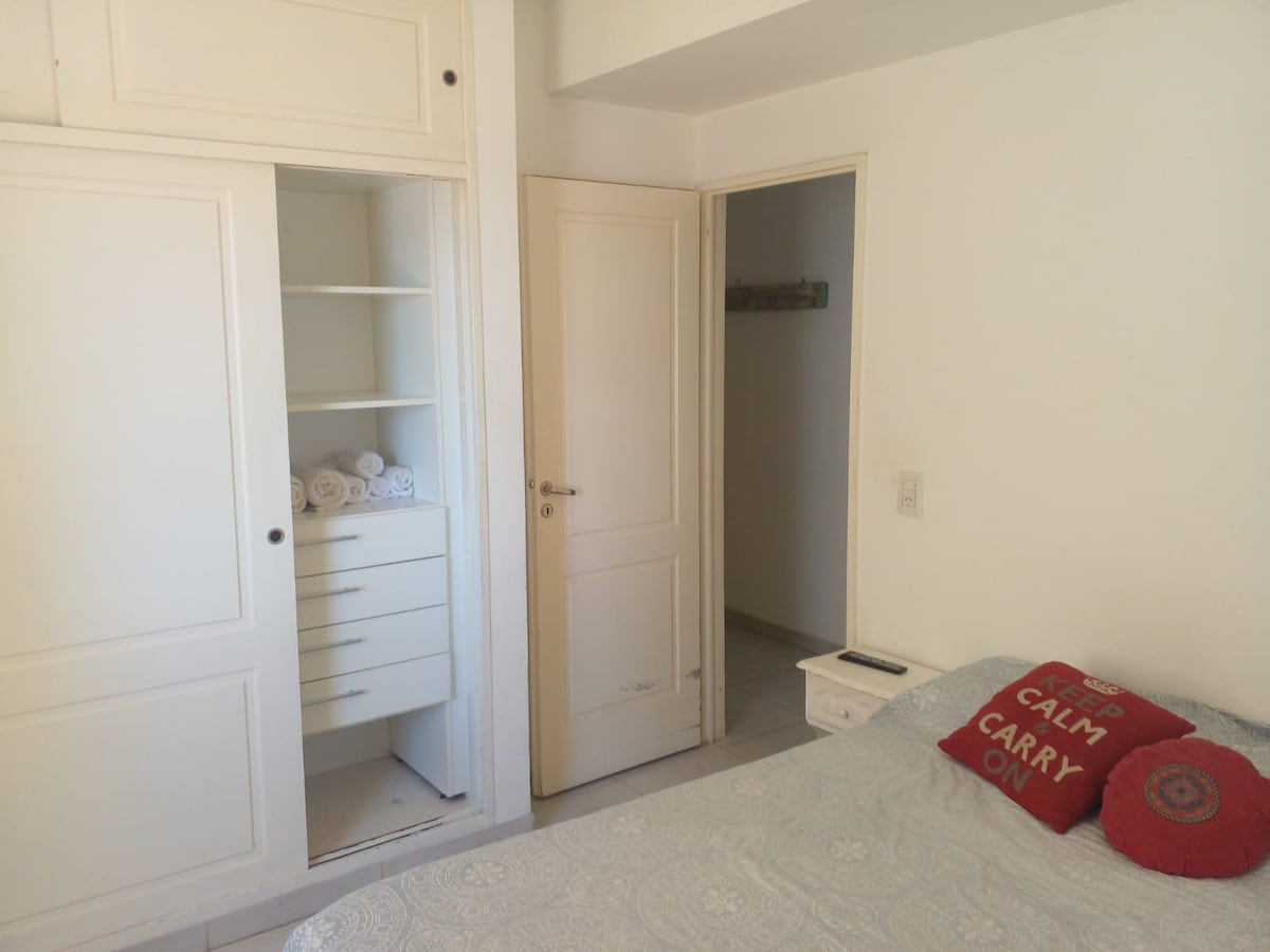 A bedroom features a spacious bed with a light-colored coverlet, and two decorative pillows. A closet with shelving is visible, showcasing neatly rolled towels. A door leads to a hallway, while light illuminates the room through a nearby opening.