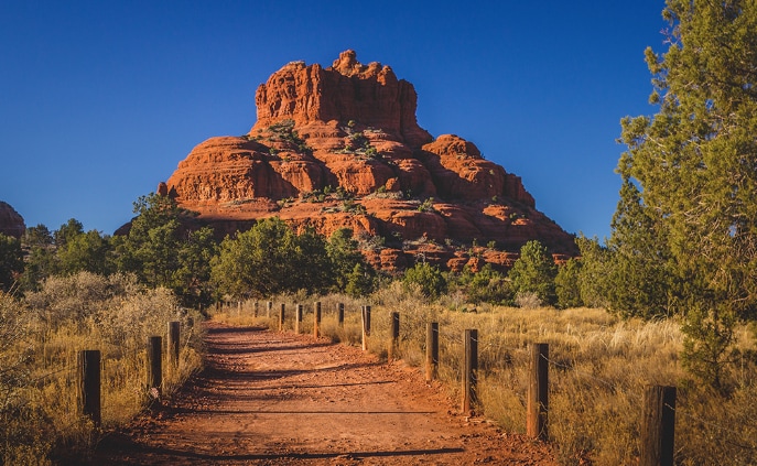 A winding dirt path approaches a prominent red rock formation surrounded by green vegetation. Wooden posts line the path, leading towards the distinctive geological features under a clear blue sky.