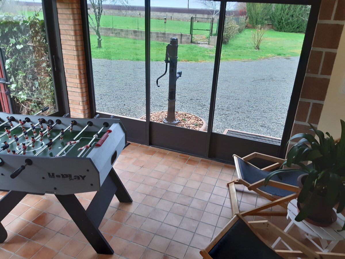 A play area combines indoor and outdoor elements, featuring a foosball table and two folding chairs positioned under large glass windows. A scenic view of a grassy area and a gravel path is visible outside, enhancing the space's connection to nature.