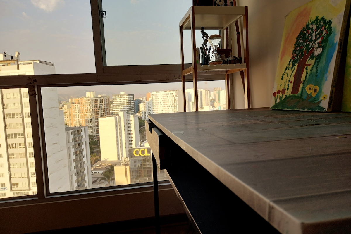 A corner workspace is illuminated by natural light from a large window. The view showcases a city skyline with various buildings. A small shelf holds decorative items, and a colorful painting of a tree is visible nearby.