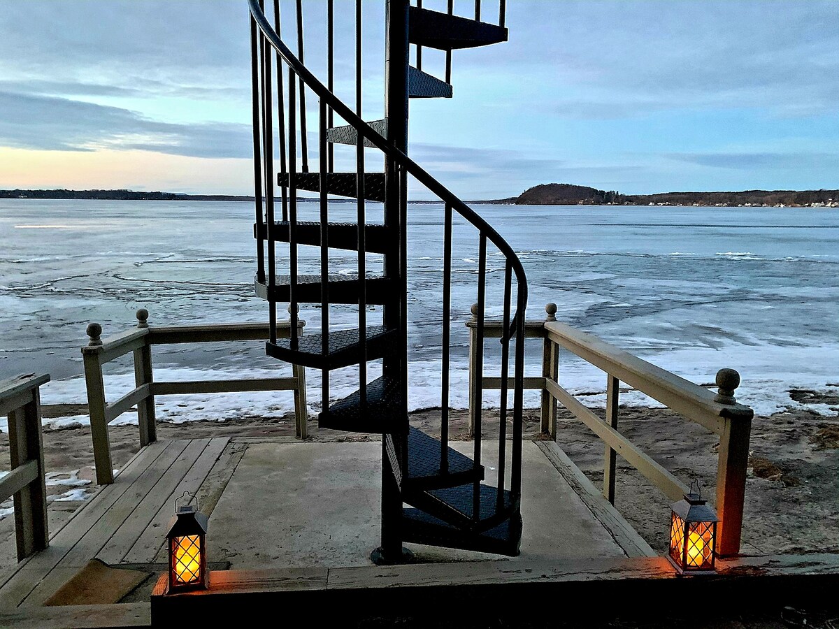 A spiral staircase leads down to a frozen lake, framed by a wooden deck. Two lanterns provide soft illumination on either side. The scene captures a serene winter landscape, showcasing ice-locked waters and a muted sky.