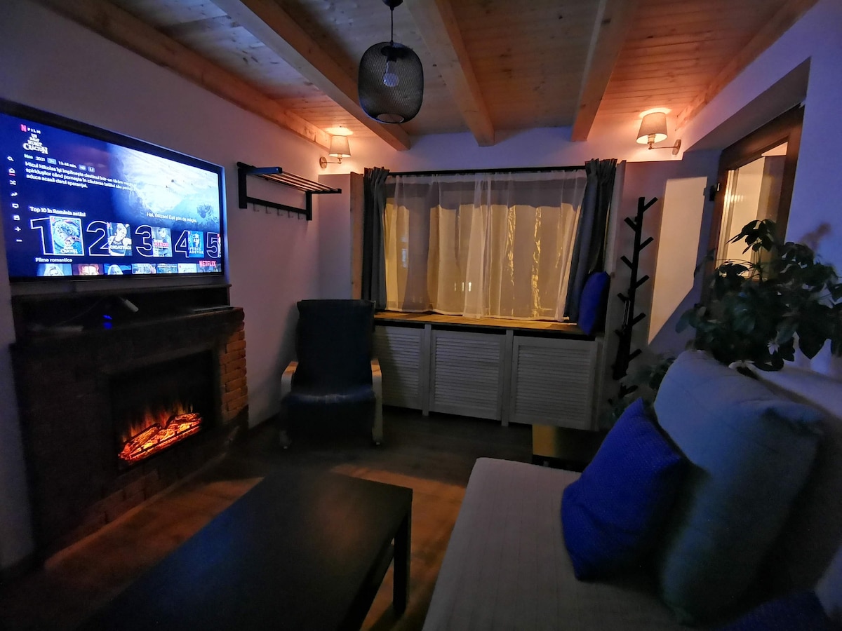 A cozy living area features a modern television mounted above a decorative fireplace. A comfortable chair and a spacious sofa with soft blue accents are positioned around a dark coffee table. Natural light filters through sheer curtains, providing a warm glow to the space.