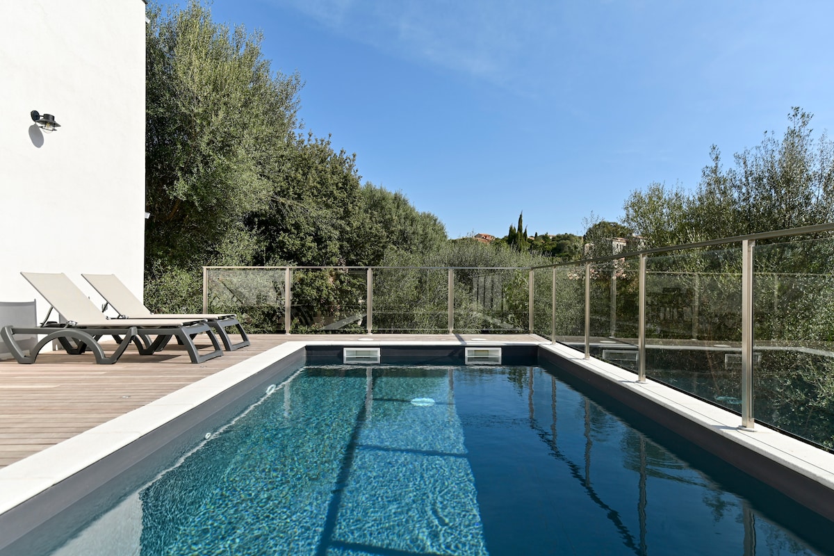 A secure swimming pool is surrounded by a glass railing, providing unobstructed views of the lush greenery. Two lounge chairs are positioned on the wooden deck, inviting relaxation by the water. The clear blue water reflects the bright sky overhead.