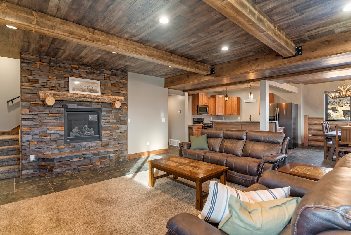 A spacious family room is highlighted by a stone fireplace, surrounded by comfortable leather couches. Reclaimed wood beams stretch across the ceiling, and a warm atmosphere is created by recessed lighting. The open design leads to a dining area and fully equipped kitchen.