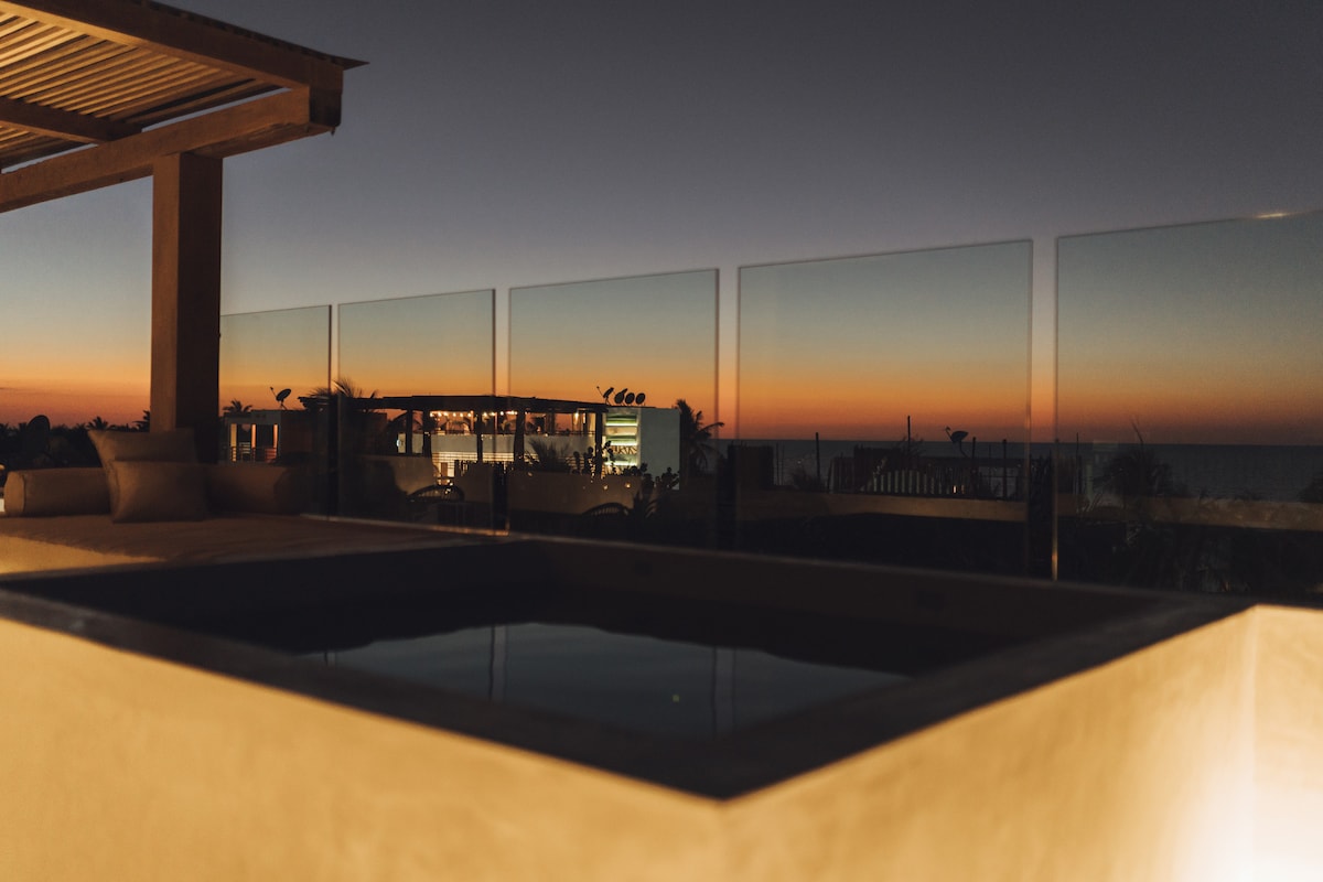A jacuzzi is illuminated softly against a backdrop of an evening sky, showcasing hues of orange and blue. Glass panels provide unobstructed views of the ocean and surrounding area, enhancing the intimate atmosphere of the outdoor space.
