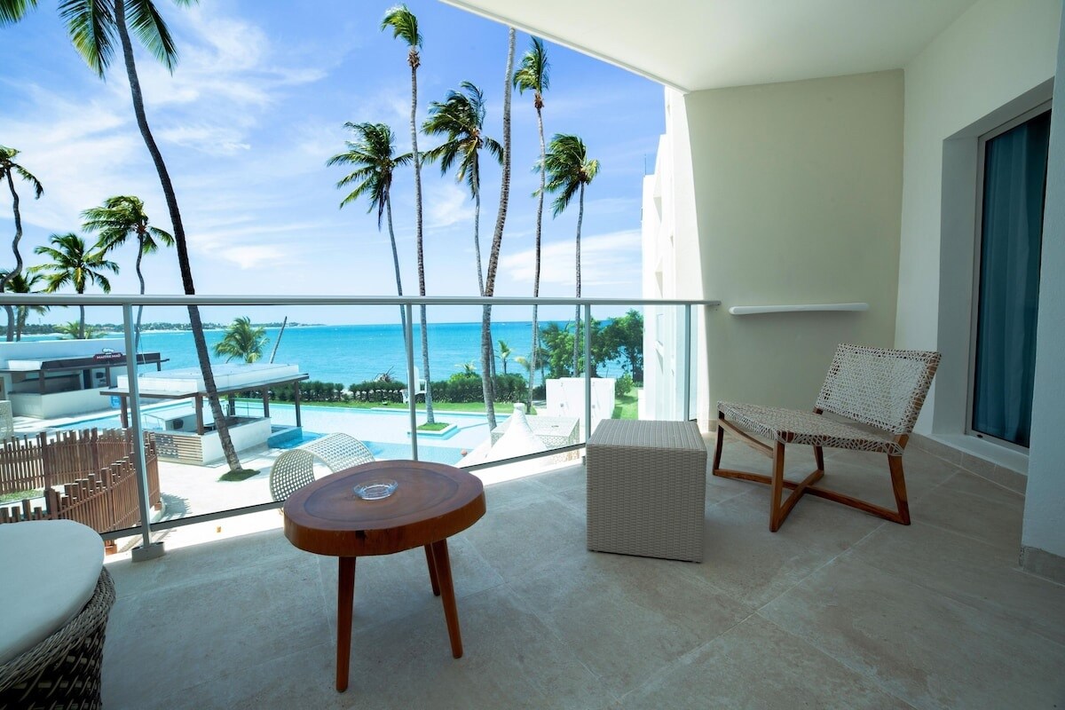 A private balcony features a wooden table and a small light-colored ottoman, complemented by a woven chair. Lush palm trees sway in the backdrop, with a glimpse of the turquoise sea and a pool visible below.