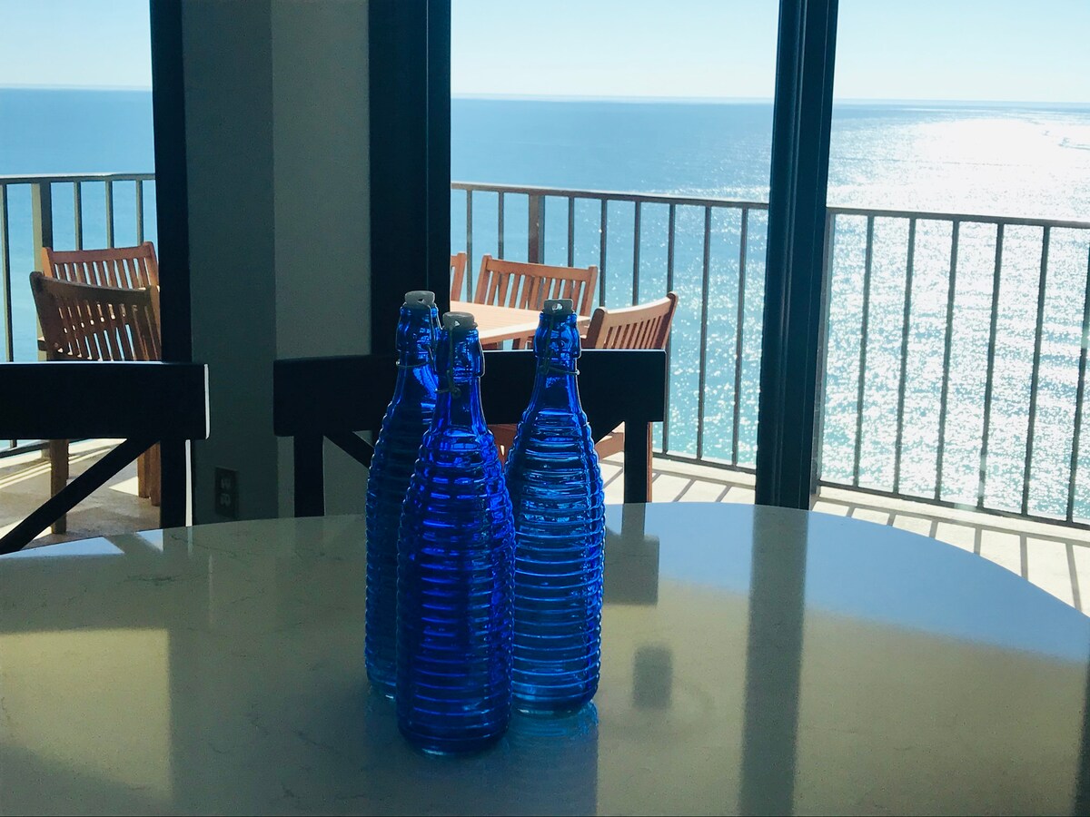 Blue glass bottles are positioned on a granite countertop, reflecting the natural light streaming through the floor-to-ceiling windows. The ocean is visible in the background, with the sun glinting off the water's surface. A wooden dining table and chairs are seen beyond the bottles.
