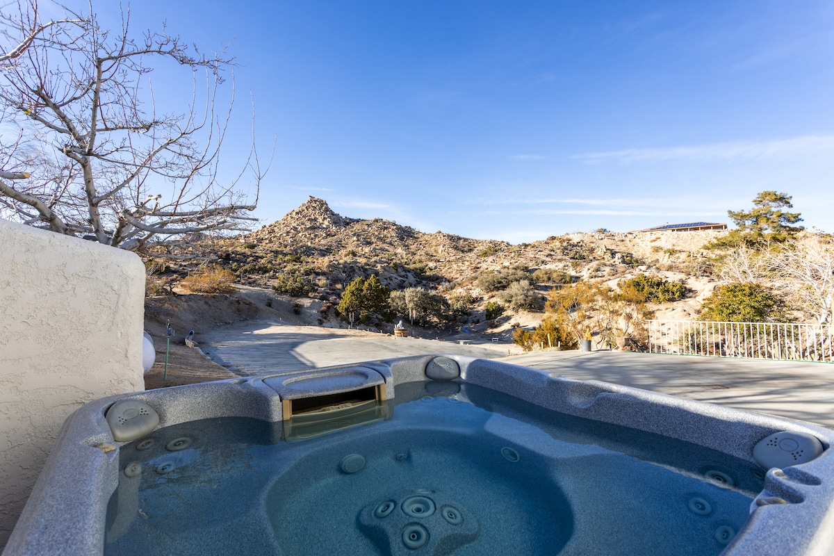 A spacious hot tub is situated outdoors, surrounded by desert landscapes. The blue waters of the tub contrast with the rocky terrain and natural vegetation in the background, with distant hills elegantly rising against a clear sky.