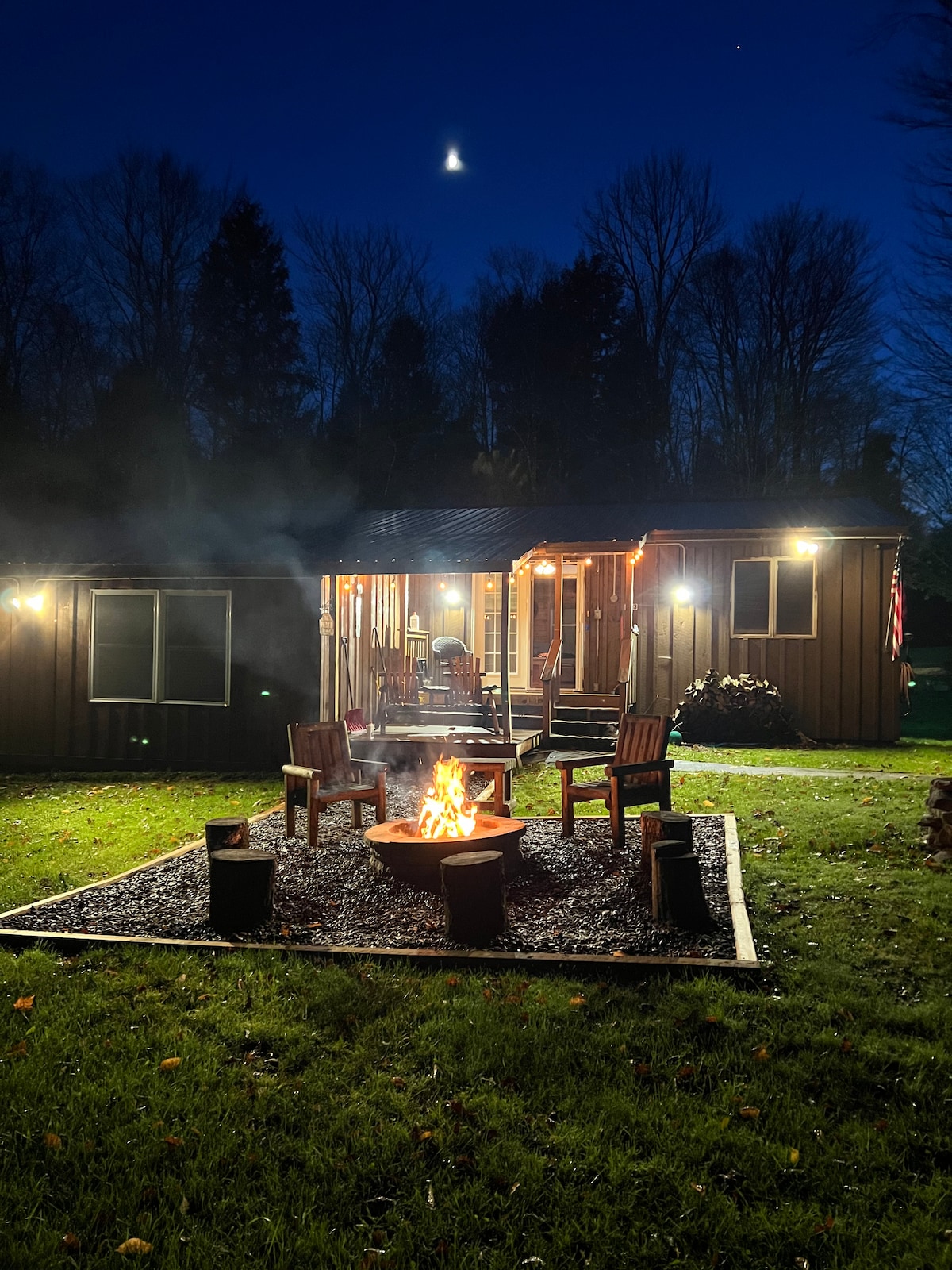 The exterior of the Cattail Manor is illuminated at night with soft lights, featuring a fire pit surrounded by several chairs. Smoke gently rises from the fire, with the deck visible in the background, providing a cozy gathering space under a crescent moon.