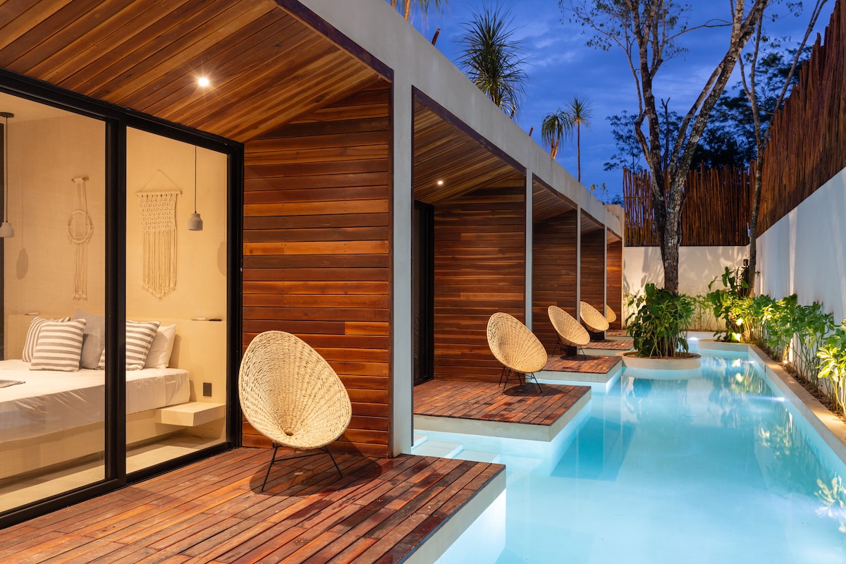 A serene outdoor space features a sparkling private pool surrounded by wooden decking. Adjacent to the pool, comfortable seating areas are framed by lush greenery and illuminated by soft evening light. The glass doors of the bedrooms provide views into the inviting interior.