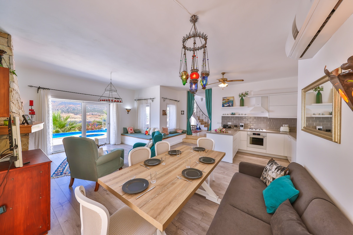 An inviting living space features a neutral color palette complemented by natural wood accents. A dining table is set for six, and a comfortable seating area includes plush cushions. Bright sunlight streams through large windows, illuminating the open kitchen area equipped with modern appliances.