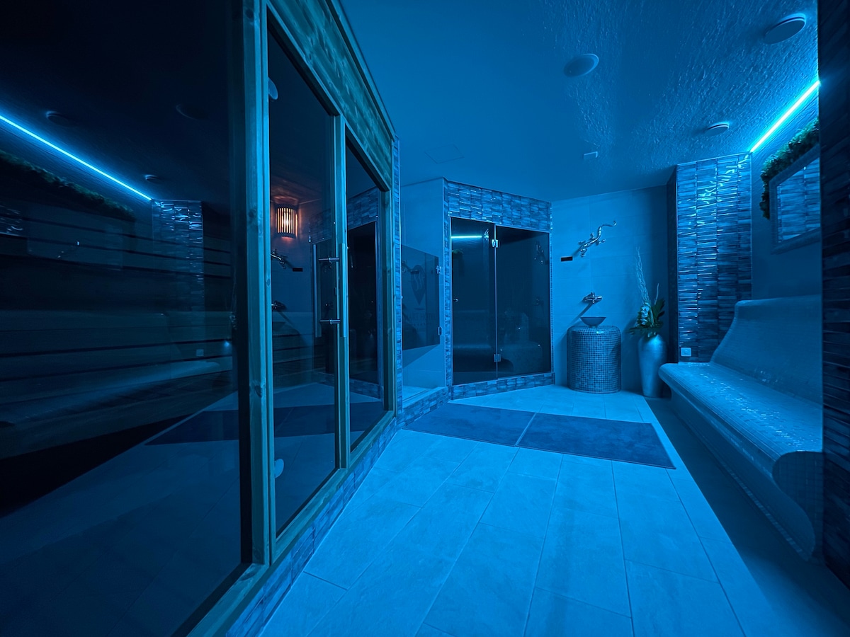 An indoor wellness area is featured, illuminated by soft blue lighting. A glass sauna is visible alongside a steam room, both designed with wood accents. A relaxation area includes a smooth stone bench, enhancing the tranquil environment for guests seeking relaxation.