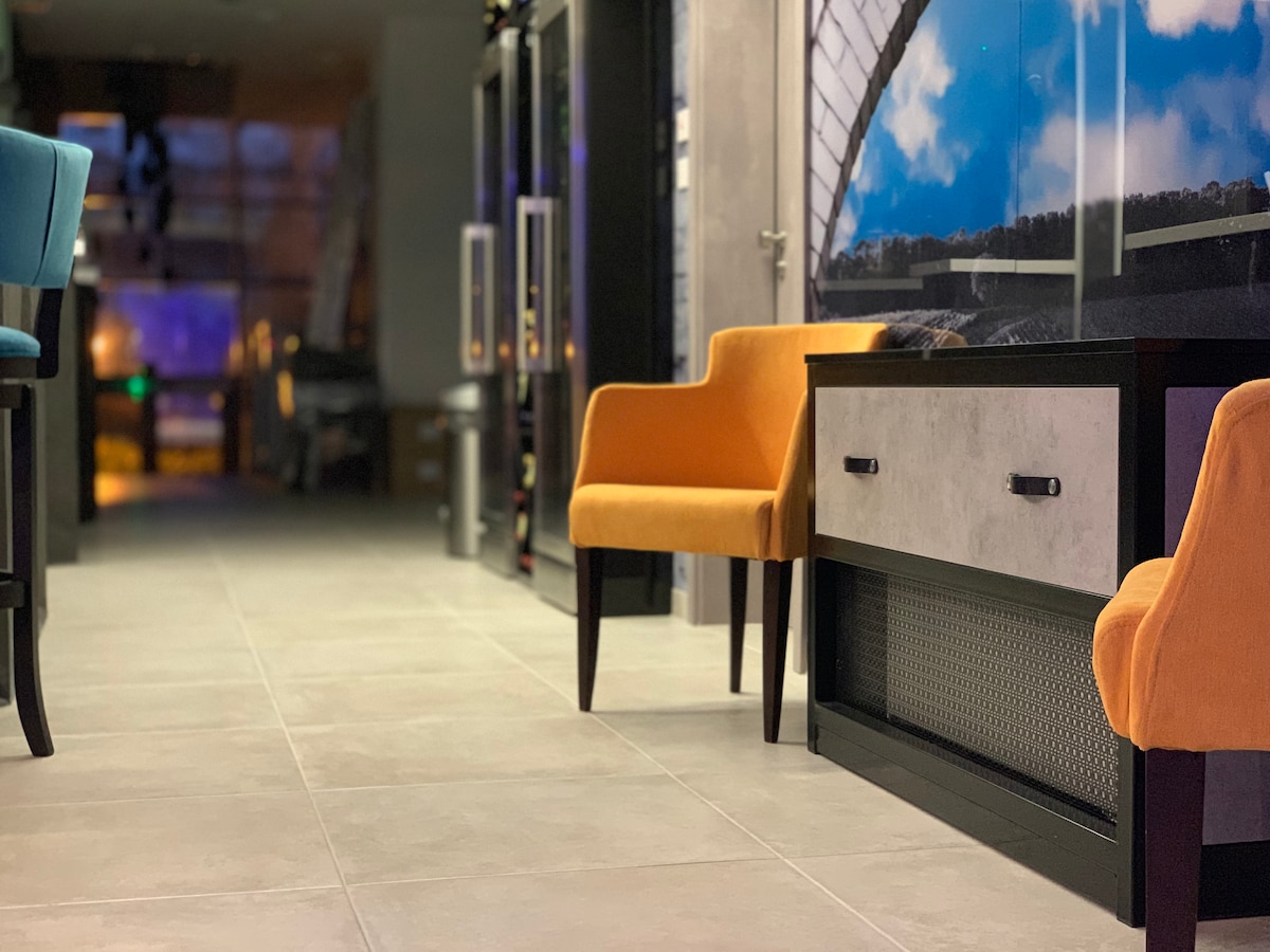 A hallway featuring tiled flooring and contemporary decor. Brightly colored chairs in blue and orange are arranged near a modern console table. The walls are adorned with large windows, allowing natural light to enhance the inviting ambiance.