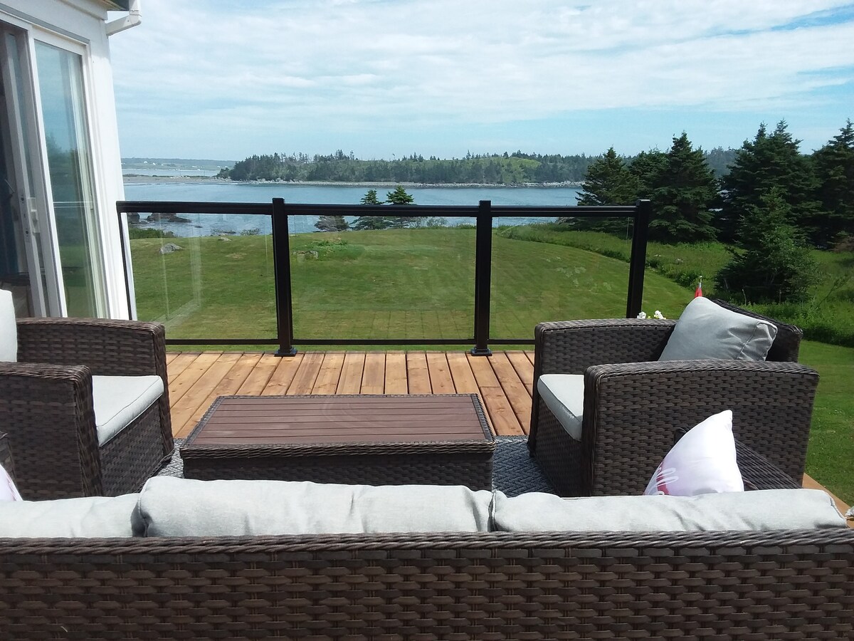 A cozy outdoor seating area is visible on a wooden deck, featuring comfortable wicker sofas with soft cushions. A glass railing offers an unobstructed view of the lush green landscape and the ocean in the background, under a clear blue sky.