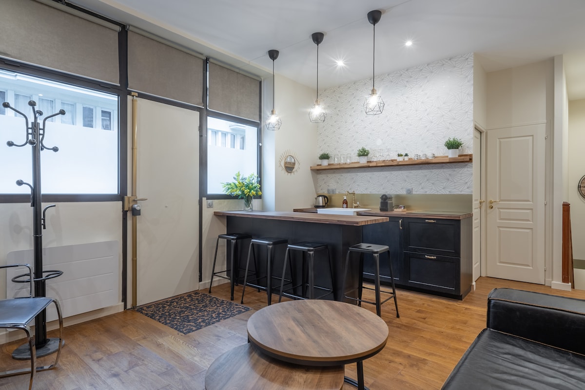A modern living area features an open kitchen with a countertop and four bar stools. Natural light enters through large windows, illuminating the wooden flooring. A circular coffee table and a coat rack add functionality to the space, which is decorated with minimalistic shelving and plants.