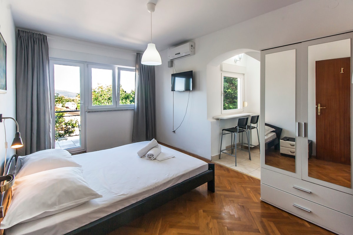 A bright studio apartment features a double bed with white linens and pillows. The space includes air conditioning, a flat-screen TV mounted on the wall, and a kitchenette with bar stools. Large windows allow natural light to fill the room, offering views of greenery outside.