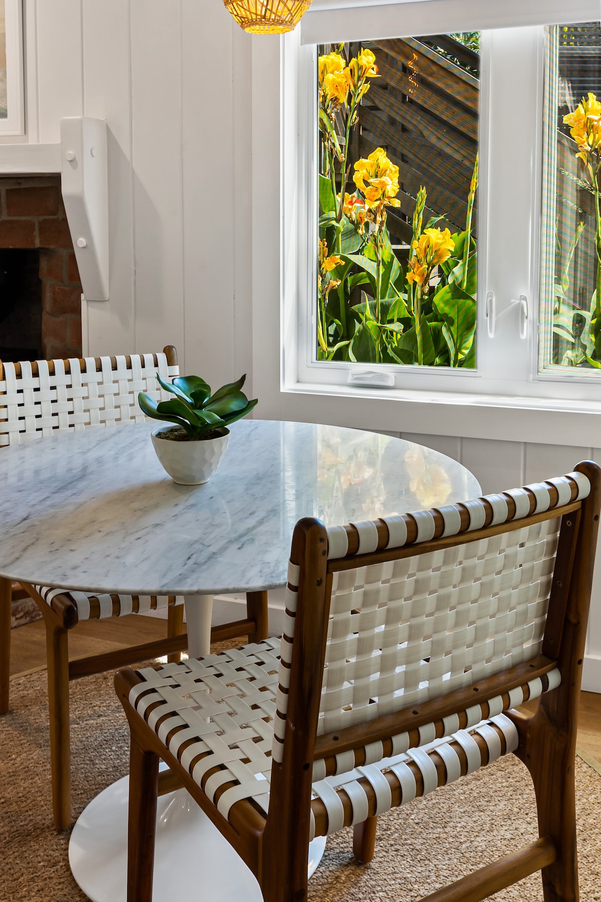 A round marble table is set with a small potted plant, surrounded by two woven chairs. Bright yellow flowers are visible through the window, offering a glimpse of nature just outside.