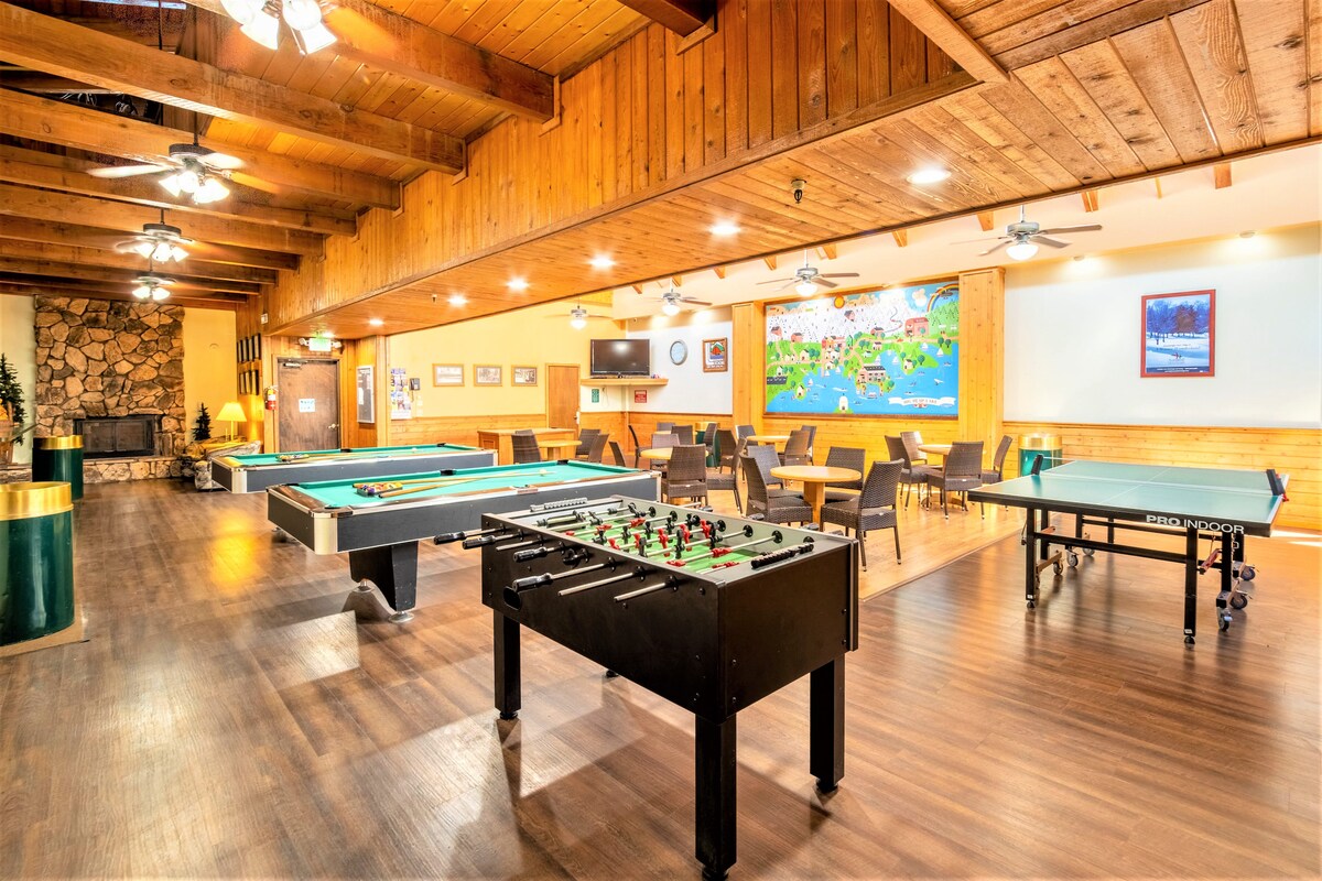 A spacious recreation room is designed for leisure activities, featuring a pool table, foosball table, and ping pong table. Wooden beams adorn the ceiling, while multiple seating areas accommodate groups. A stone fireplace adds warmth, contributing to a cozy atmosphere.