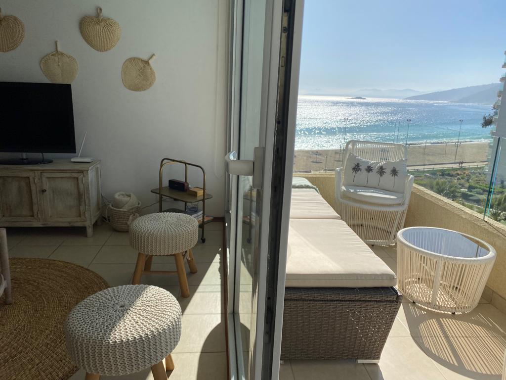 A bright living area opens to a balcony, featuring a cozy seating area with woven stools and a daybed. The serene ocean view is visible through large glass doors, inviting natural light into the space. Decorative straw wall hangings add a hint of texture.