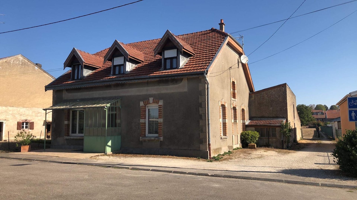 The exterior of an old café is depicted, featuring a mix of brick and stone walls, a tiled roof, and multiple windows. A small front porch is visible, with greenery placed near the entrance. The surrounding area includes gravel paths and nearby structures, indicating a quiet street setting.