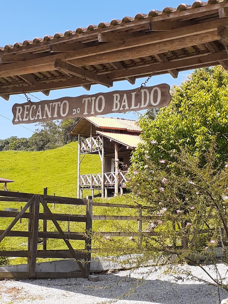 A wooden sign reading 'Recanto do Tio Baldo' hangs above a rustic entrance. In the background, a two-story house is visible, surrounded by lush greenery and hills. The setting conveys a serene atmosphere, inviting guests to explore the natural surroundings.