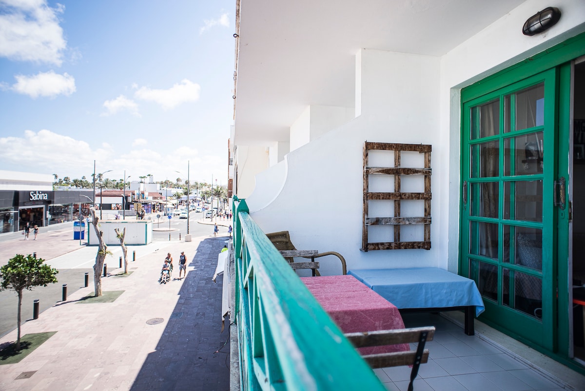 A balcony is featured, offering views of the vibrant street below. The green railing complements the light-colored walls. A small table and two chairs are visible, creating a comfortable space for relaxation. Sunlight illuminates the area, enhancing the lively outdoor atmosphere.