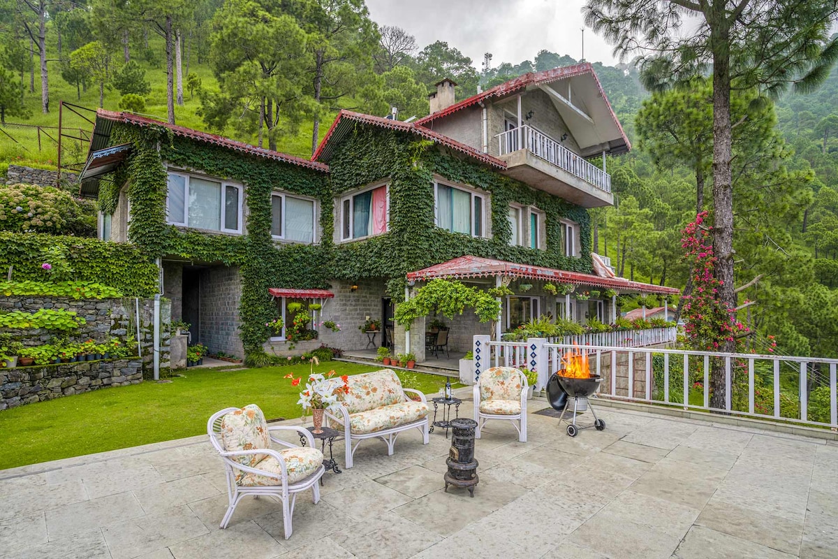 A charming facade of the villa is surrounded by lush greenery, with creeping vines adorning the stone-clad exterior. An inviting patio features comfortable seating arrangements, alongside a barbecue grill, all set against the backdrop of mountainous terrain and a well-maintained lawn.
