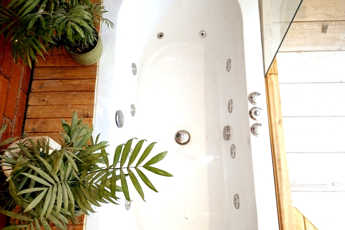 A spacious bathtub is positioned against a wooden deck, surrounded by potted green plants. The tub features multiple jets along its sides, providing a modern touch. Natural light is captured reflecting off the smooth, white surface.