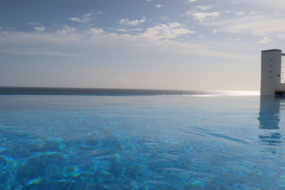 An infinity pool stretches out towards the ocean, blending seamlessly with the water. The surface reflects the sky and shimmering sunlight, creating a serene ambiance. The horizon is visible, meeting the sky in a soft gradient, enhancing the tranquil setting.