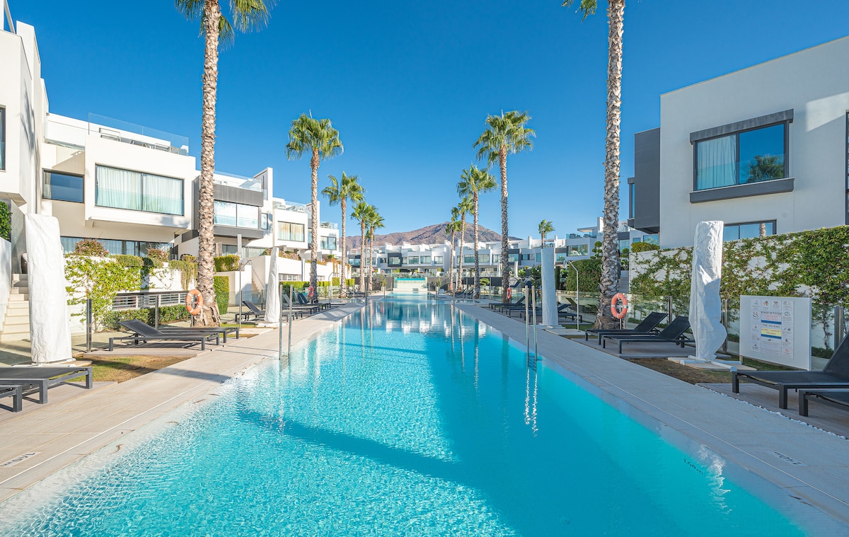 A long, crystal-clear swimming pool stretches between modern townhouses surrounded by palm trees. Loungers line the pool's edge, providing ample space for relaxation. Sunlight reflects off the water, creating a serene atmosphere, complemented by the well-kept landscaping along the pathway.