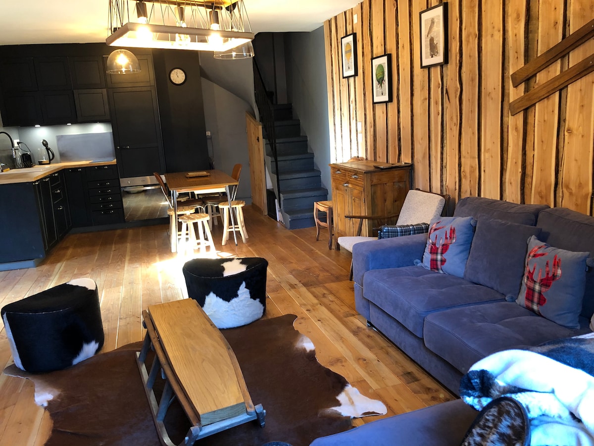 The living area features wooden wall paneling and a comfortable seating arrangement, including a spacious blue sofa and cowhide poufs. A wooden coffee table is centered on a patterned rug. An open kitchen and dining area, equipped with a table and chairs, is visible in the background.